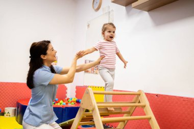 Pediyatrik fizikçi hevesli bir çocuğa evde tahta merdiven tırmanma, motor beceri geliştirme ve terapi seansı sırasında fiziksel rehabilitasyonu teşvik etme konusunda yardımcı oluyor.