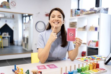 Konuşma terapisti kadın alfabe bilgi kartları ve renkli oyuncaklarla canlı bir konuşma terapisi yapıyor, anaokulu çocuklarına fonetik, eklem ve dil becerileri öğretiyor.