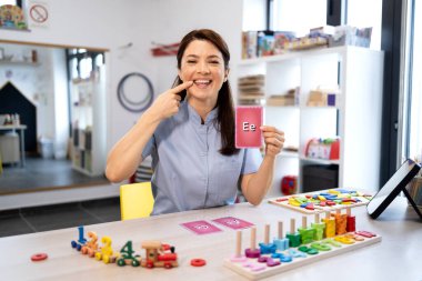 Konuşma terapisti düzgün ağız oluşumunu gösteriyor ve bir dil terapisi seansında alfabe bellek kartı tutuyor. Klinik ortamında konuşma geliştirme konusunda yardımcı oluyor.