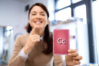 Konuşma terapisti elinde C harfiyle çenesine işaret eden bir hafıza kartı tutuyor. Klinikte veya eğitim ortamında verilen özel bir ders sırasında fonetik ve konuşma geliştirme egzersizlerine odaklanmak