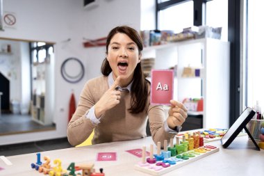 Konuşma terapisti, ses üretimi ve harf tanıma programını, destekleyici bir öğrenme ortamında iletişim yardımı ve erken eğitim sunan bir kartla göstermektedir.