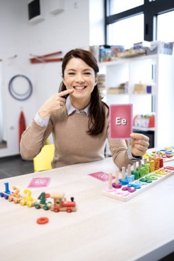 Konuşma terapisti gülümsüyor, elinde bir alfabe kartı tutarken ağzını işaret ediyor. Eğitimsel ahşap oyuncaklarla yapılan bir dil terapisi sırasında fonetik ve doğru ifade etmeyi öğretiyor.