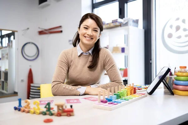 Genç bir kadın konuşma terapisti parlak bir klinikte kameraya gülümsüyor, ahşap sayma treni, alfabe kartları ve oyuncaklarla dolu bir masada oturuyor, öğrenme için sıcak profesyonel bakım sunuyor.