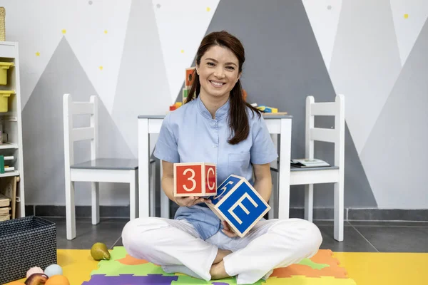 Konuşma terapisti çocuk dostu bir sınıfta renkli bir oyun mat üzerinde oturuyor, rakamlar ve harflerle ahşap alfabe blokları tutuyor, erken eğitim ve iletişim terapisine odaklanıyor.
