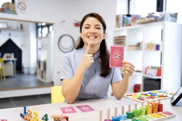 Konuşma terapisti kadın alfabe bilgi kartları ve renkli oyuncaklarla canlı bir konuşma terapisi yapıyor, anaokulu çocuklarına fonetik, eklem ve dil becerileri öğretiyor.