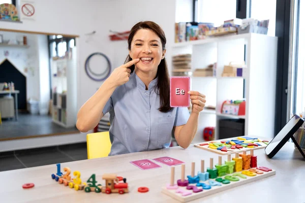 Konuşma terapisti düzgün ağız oluşumunu gösteriyor ve bir dil terapisi seansında alfabe bellek kartı tutuyor. Klinik ortamında konuşma geliştirme konusunda yardımcı oluyor.