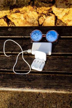 Güneş gözlüğü, akıllı telefon, elektrik bankasından şarj edilen kablosuz kulaklıklı bir kutu, ahşap bir bankta yatıyor.