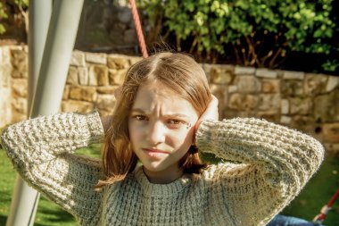 Güzel genç kız kulaklarıyla kulaklarını kapatıyor çünkü kulakları ağrıyor ve güneşli bir günde şehirdeki oyun alanındaki gürültüyü duymak istemiyor.