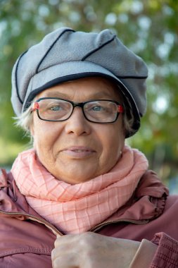 Güneşli bir günde mavi gökyüzüne karşı gri bereli ve pembe eşarplı yaşlı bir kadının portresi.