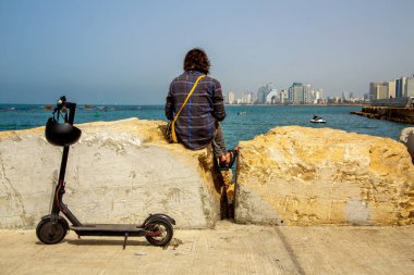 Ekose gömlekli genç bir adam taşların üzerinde oturuyor ve kasklı elektrikli scooterın arkasındaki Tel Aviv şehrine bakıyor. Parlak güneşli bir gün
