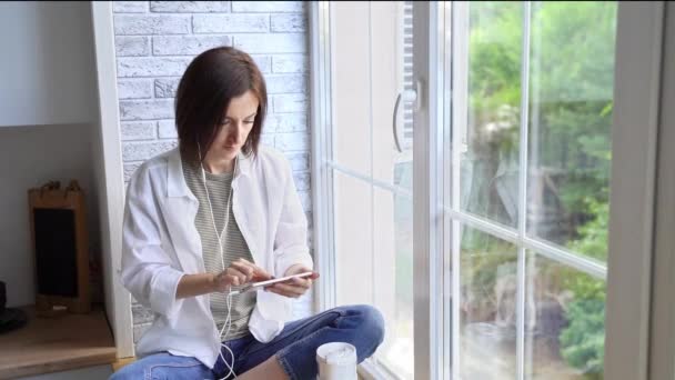 창문에 앉아 있는 젊은 여자. 휴대폰으로 일하고 있습니다. 온라인 연구 개념 — 비디오