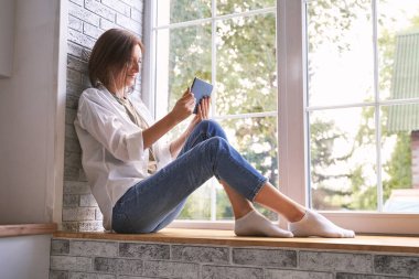 Pencere kenarında oturan genç bir kadın. Tablet üzerinde çalışıyorum. Çevrimiçi çalışma kavramı.