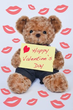 A cute teddy bear holding a yellow sign that says Happy Valentine's Day surrounded by kisses on a white background