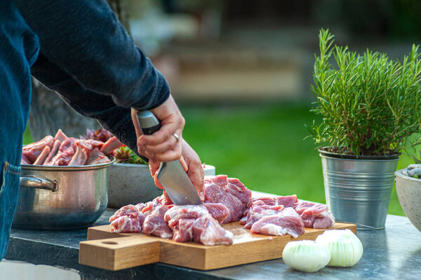 Preparation for BBQ: man's hand slicing meat and onion with big knife on wooden cut board, outdoors