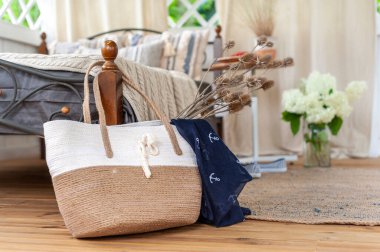 Apartmanın yatak odasındaki yatağın yanında açık renk bir arka planda büyük kadınların hasır çantasıyla.