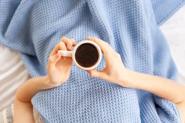 a cup of coffee or tea in the gentle hands of a young girl lying in bed under a blanket, top view