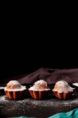 Siyah arkaplanda peçeteler olan tahtanın üzerine pudra şekeri serpiştirilmiş kapkekler.