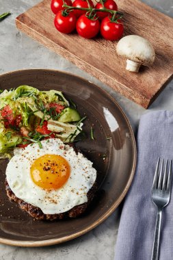 Çırpılmış yumurta ve salata kahverengi bir tabakta, üzerinde domatesler ve gri beton arka planda mantarlar olan tahtanın yanında.
