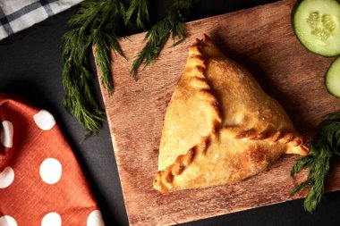 Ahşap bir tahtanın üzerinde üç köşeli turta, salatalıklı, dereotlu ve siyah arka planda peçeteli.