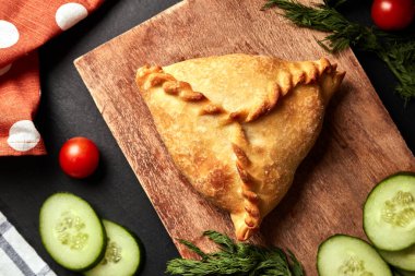 Ahşap bir tahtanın üzerinde üç köşeli turta, salatalıklı, dereotlu ve siyah arka planda peçeteli.