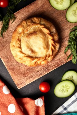 Ahşap bir tahtanın üzerinde yuvarlak turta, salatalıklar, domatesler, otlar ve siyah arka plan manzaralı bir peçete.