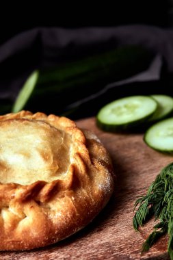 Tahtada yuvarlak pişmiş turta, salatalıklı, siyah arka planda otlar.