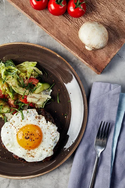 Çırpılmış yumurta ve salata kahverengi bir tabakta, üzerinde domates ve mantar olan ahşap bir tahtanın yanında gri beton arka plan manzaralı.