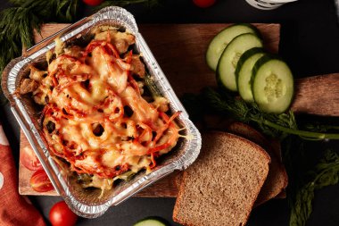 Tahta bir tahtanın üzerindeki termal konteynırda lazanya, salatalık ekmeği ve siyah arka plan manzaralı peçete.