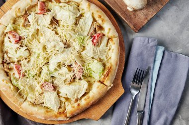 Tahtada pizza, gri beton arka planda, peçeteler ve çatal bıçaklar üst tarafta.