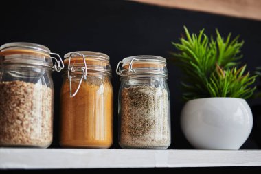 Tahıllı cam kavanozlar ve siyah arka planda beyaz bir rafta dekoratif bir çiçek.