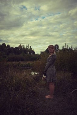 Elinde sepeti olan köylü elbiseli sarışın bir kız ve kasvetli bir tarlada küçük bir köpek.