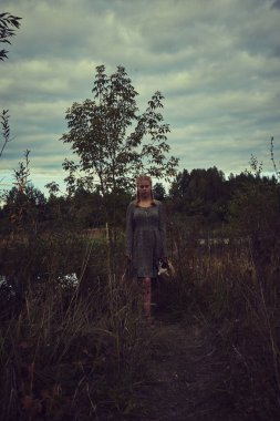 Elinde sepeti olan köylü elbiseli sarışın bir kız ve kasvetli bir tarlada küçük bir köpek.