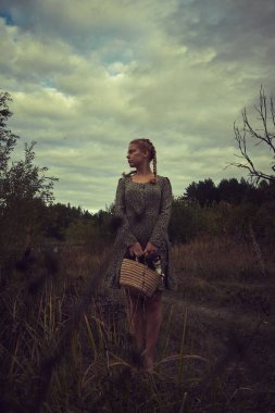 Elinde sepeti olan köylü elbiseli sarışın bir kız ve kasvetli bir tarlada küçük bir köpek.