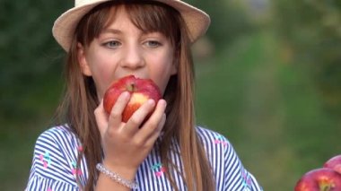 Güneşli Meyve Bahçesinde Kırmızı Elma Yiyen Güzel Mavi Gözlü Kız. Güzel Gözlü Gülümseyen Küçük Kız Kameraya Bakıyor. Sağlıklı gıda konsepti. Şapkalı ve Taze Elmalı Şirin Kız Fotoğrafı.