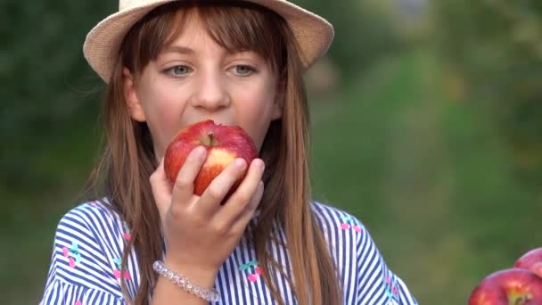 在阳光花园吃红苹果的漂亮的小蓝边女孩 笑着的小女孩美丽的眼睛看着相机 健康食品概念 用帽子和新鲜苹果近距离拍摄可爱女孩 — 图库视频影像