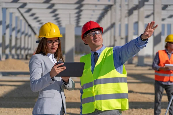 Businessman and Young Female Architect With Digital Tablet Meeting at Construction Site. Businessperson Pointing at Industrial Building Under Construction. Gender Equality at Work.