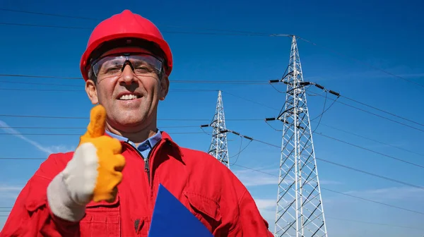 Gülümseyen Elektrik Hizmetleri İşçisi Elektrik Aktarım Kulelerinin yanında Başparmak Veriyor. Kırmızı Hardhat ve Kapaklı Mühendis Portresi. Bir Yüksek Voltajlı Elektrik Aktarım Kuleleri Hattı.