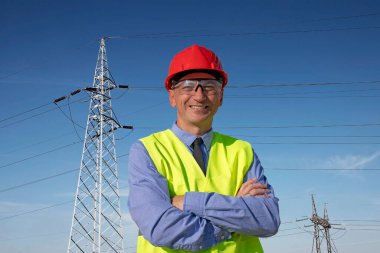 Kırmızı Şapkalı Gülümseyen Mühendis ve Elektrik Aktarım Kulelerinin Altında Duran Sarı Batı. Kırmızı şapkalı, mavi gömlekli ve sarı yelekli Happy Manager kameraya bakıyor. İnşaat Ustabaşı, Denetmen, Mühendis veya İşadamı Portresi. 
