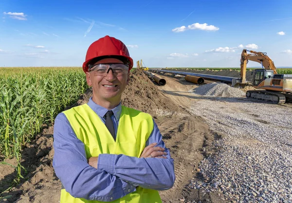 Smiling Construction Supervisor in Red Hardhat Standing at Gas Pipeline Construction Site - Turkish Stream. Happy Manager in Red Hardhat, Blue Shirt and Yellow Vest Looking at Camera.
