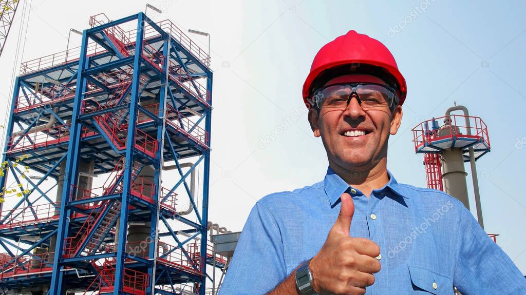 Smiling Petroleum Engineer Standing in front of Oil and Gas Refinery. Happy Oil Worker in Red Hardhat Giving Thumb Up. Portrait Of Smiling Man in Protective Workwear.