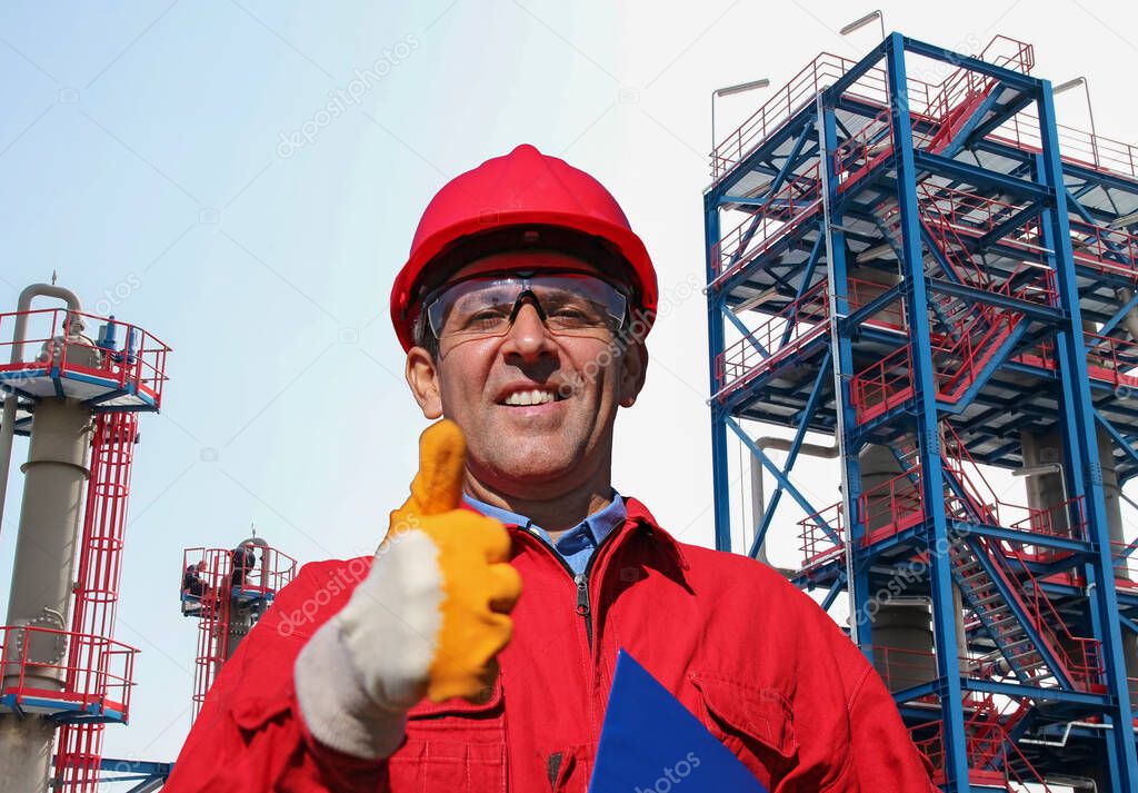 Oil and Gas Refinery Worker in Red Hardhat and Overalls Looking At Camera. Happy Oil Worker Giving Thumb Up in front of Natural Gas Processing Plant. Portrait Of Smiling Man in Protective Workwear.