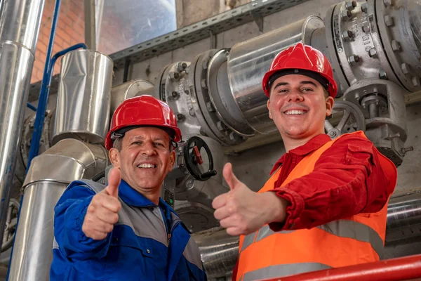 Smiling Petroleum Engineer and Young Worker in Red Coveralls and Hardhat Giving Thumbs Up Beside Industrial Piping. Two Oil Refinery Workers in Personal Protective Equipment Standing Inside Refinery.