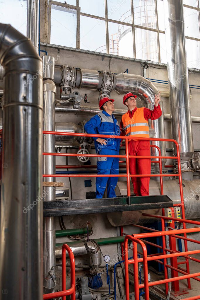Maintenance Engineer Standing Next to Oil Worker in Oil Refinery. Oil Refinery Workers in Personal Protective Equipment Talking in Industrial Interior With Large Piping. 