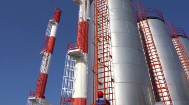 Enerji Santrali İşçisi, endüstriyel bir deponun dış cephesindeki Metal Merdivenden Tırmanıyor - Yakınlaş. Endüstriyel bacaları, depolama tankı ve Mavi Gök 'e karşı çalışan işçisi olan bir elektrik santrali..