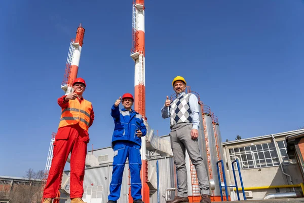 Manager, Engineer and Power Plant Worker Giving Thumbs Up in front of Industrial Chimneys. Two Workers in Personal Protective Equipment Meeting with Businessperson. Success and Teamwork Concept.