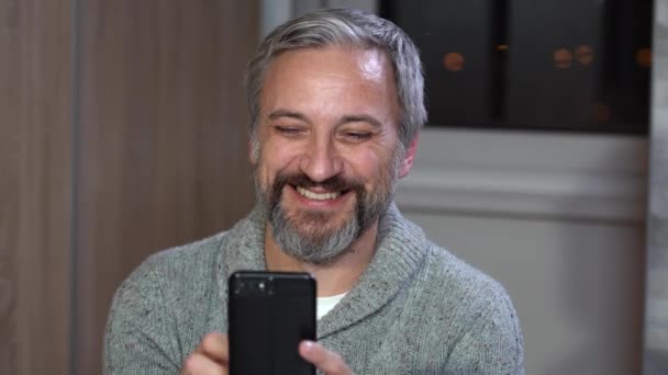英俊的灰头发男人拿着智能手机 看着相机 笑着的肖像 快乐微笑的成年人在家里用手机 — 图库视频影像