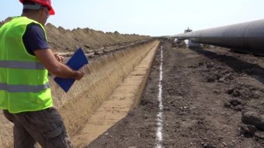 İnşaat Ustası Gaz Boru Hattı İnşaat Alanında İlerleme Kontrol Ediyor. Tahtaya Denetmen Yazıyor. Kırmızı Şapkalı ve Sarı Yelekli Mühendis Portresi.