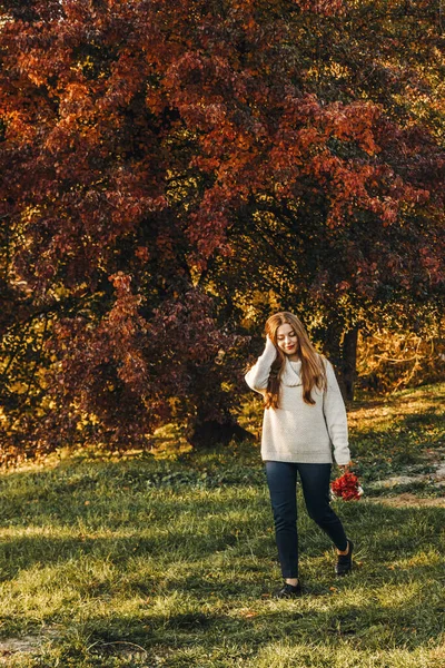 Genç kadın sonbaharda parkta yürüyor. Onun elinde kırmızı çiçekler var. O bej bir kazak giyiyor ve saçı uzun..