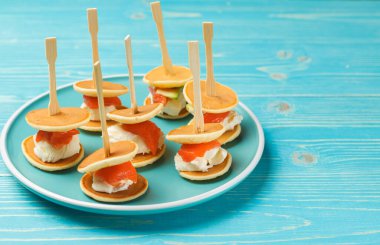 Parti ve resepsiyonlar için orjinal bir atıştırmalık. Mini krep, yumuşak peynir ve somon. Mavi ahşap arka planda mini etiketleri olan bir tabak. Yatay çerçeve, boşluğun kopyası