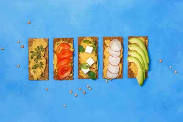 健康的でベジタリアンスナックのコンセプト フムス トマト アボカド 種子でライ麦トーストにサンドイッチ 明るい青の背景に ハードライト コピースペースのオーバーヘッド — ストック写真